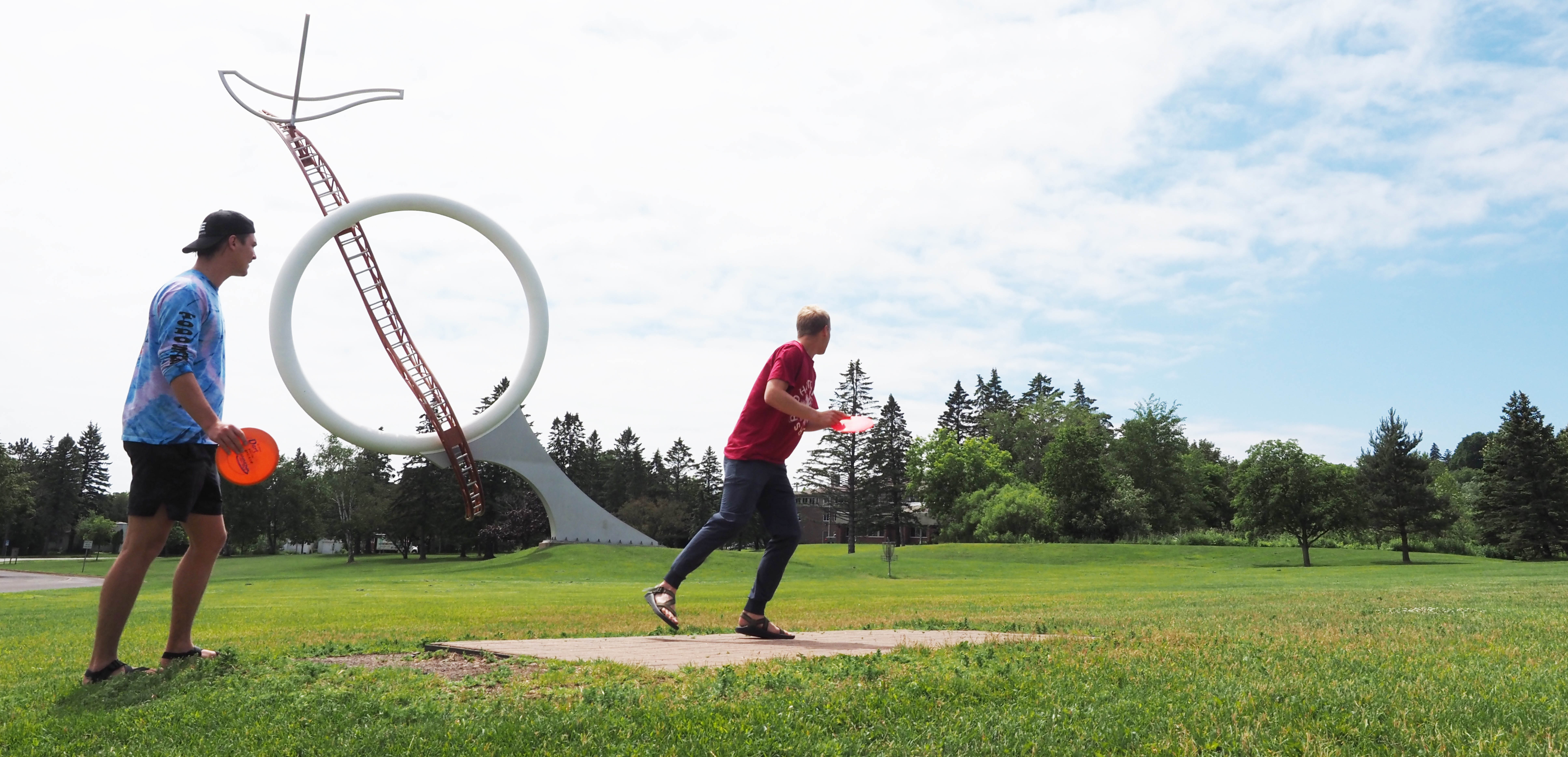 two students disc golfing by the ricing moon sculpture